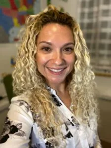 Smiling Sparrow Media team member with curly blonde hair in a white floral blouse, in a bright Central NY office